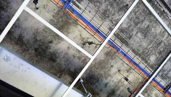 Cable tray with wires and metal duct show above t-bar grid in opened suspended (drop) ceiling and below concreate of upper floor at construction site of industrial building
