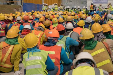 Bir grup inşaat işçisi güvenlik üniforması giyip inşaat alanında toplanıyor.