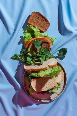 Mavi bir örtü üzerine çeşitli yeşillikler serpiştirilmiş düz bir ekmek, sağlıklı sandviç konsepti, seçici odaklanma.