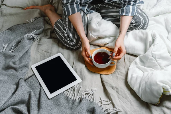 Tablet ve çayla yatağında oturan yüzsüz kadın portresi, seçici odaklanma.