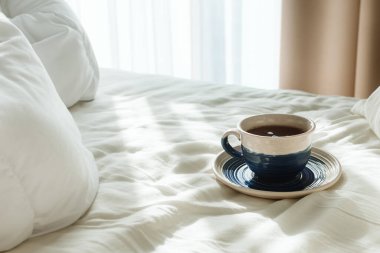Beyaz çarşaflı yatak ve yastıklı ve sabah güneşinde bir fincan sıcak çay ya da kahve, seçici odak