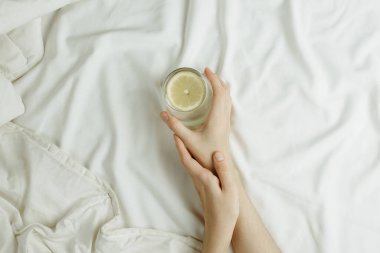 Beyaz çarşaflı yatakta limon suyunda cam tutan kadın elleri, sağlıklı bir sabah kavramı.