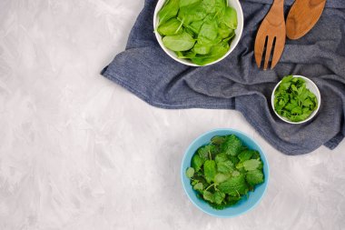 Çimento arkaplanındaki salata için taze yeşiller: Ispanak, nane ve maydanozlu lacivert havluda ahşap kaşıklar, kopyalama alanı