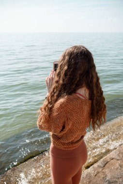 Arka plandaki videodaki kadın ya da sahilde akıllı telefonuyla resim çeken, seçici odaklı kadın.