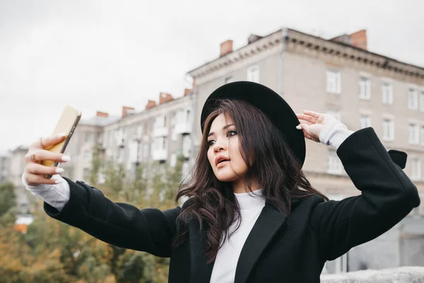Koyu renk ceketli ve şapkalı Asyalı genç bir kadın dışarıda selfie çekiyor.