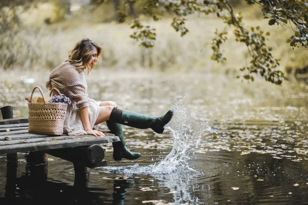 Donna con stivali di gomma seduta sul ponte di legno vicino al lago — Foto Stock
