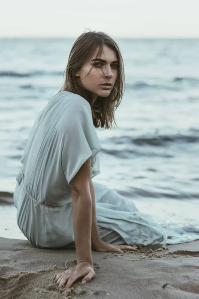 Vackra, sensuella kvinnan i en lång klänning som sitter i sanden på stranden — Stockfoto