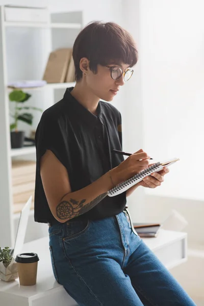 Attraktive Geschäftsfrau mit Brille sitzt am Schreibtisch im Büro und macht sich Notizen — Stockfoto