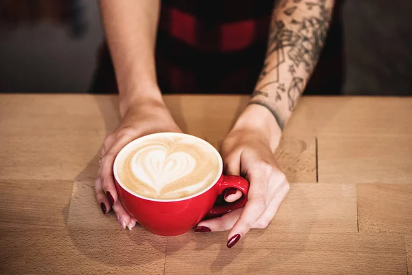 Pandangan atas tangan wanita memegang cangkir merah kopi panas berdiri di atas meja kayu — Stok Foto