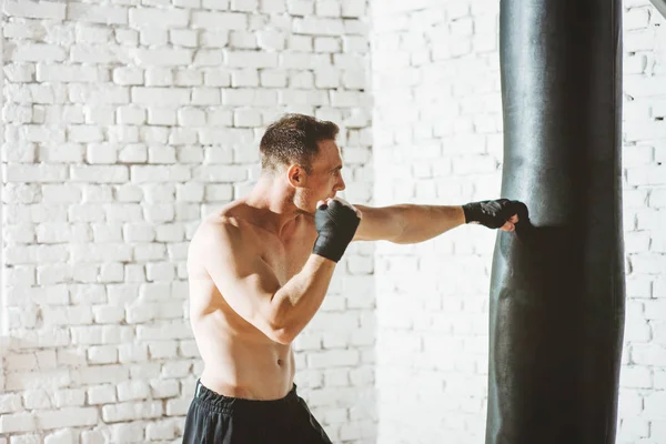 Muscular fighter practicing with punching bag against white brick wall. — Stock Photo, Image