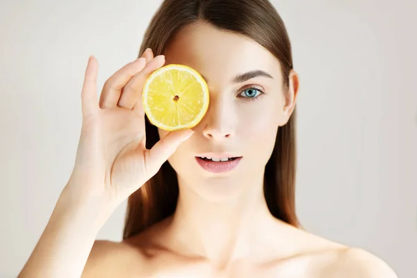 Mulher bonita com pele perfeita limpa escondendo olho atrás de fatia de limão no fundo branco . — Fotografia de Stock