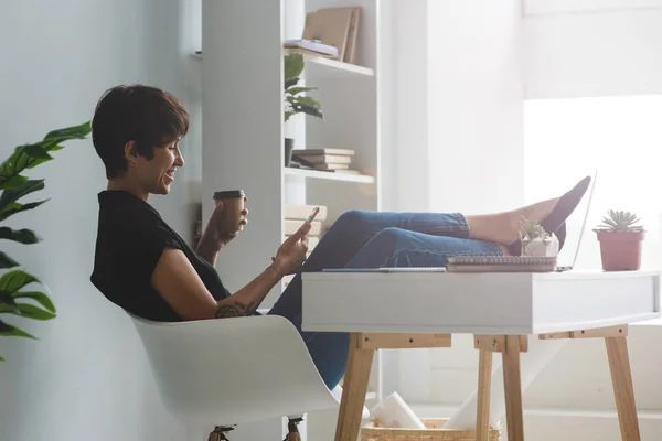 Досить молода бізнес-леді сидить у кріслі зі своїм смартфоном з ногами над столом . — стокове фото