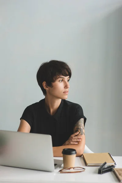 Junge Geschäftsfrau sitzt mit verschränkten Armen am Schreibtisch und schaut weg. — Stockfoto