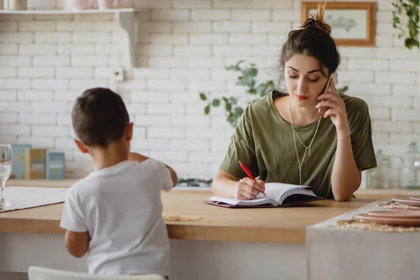 Νεαρή γυναίκα κρατά σημειώσεις και μιλάει στο τηλέφωνο, ενώ το παιδί της παίζει κοντά. — Φωτογραφία Αρχείου