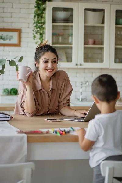Νεαρή μητέρα χρησιμοποιεί φορητό υπολογιστή ενώ παίρνει πρωινό στην κουζίνα με το μικρό της γιο. — Φωτογραφία Αρχείου