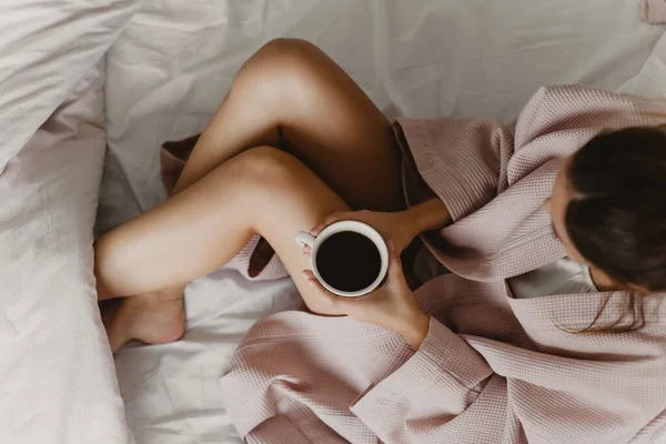 Γυναίκα σε ροζ ντουλάπα κρατώντας ένα φλιτζάνι ζεστό καφέ στο κρεβάτι. — Φωτογραφία Αρχείου