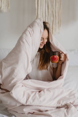 Güzel bir kadın battaniyenin altında saklanıyor ve yatakta kahve içiyor..