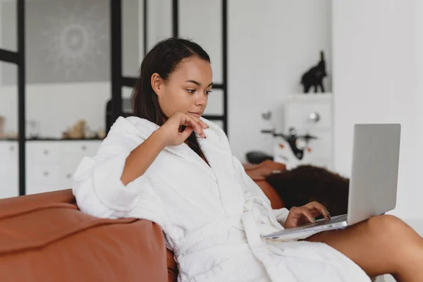 ผู้หญิงแอฟริกันอเมริกันที่น่าสนใจใช้แล็ปท็อป ขณะที่นั่งที่โซฟาที่บ้าน . — ภาพถ่ายสต็อก