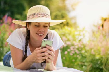 Genç neşeli kadın yaz bahçesinde dinleniyor. Akıllı telefonuyla arıyor.