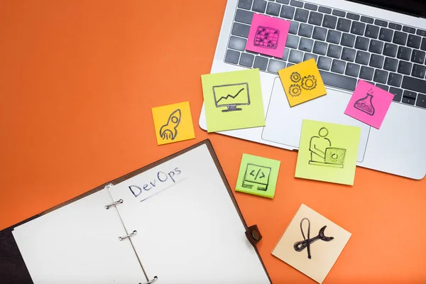 Devops konsepti. Geliştiriciler ve işlemler sözcükler ve semboller, dizüstü bilgisayar, turuncu arkaplanda boş sayfalar olan defter. Metnin için boşluk.