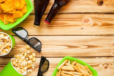 Hafta sonu evde, boş zaman tarzı, TV, fast food konsepti. 3D gözlükler, yeşil kaselerde uzaktan kumanda ve aperatifler: cips, patlamış mısır, ahşap arka planda hamburger. Üst Manzara. Kapat.