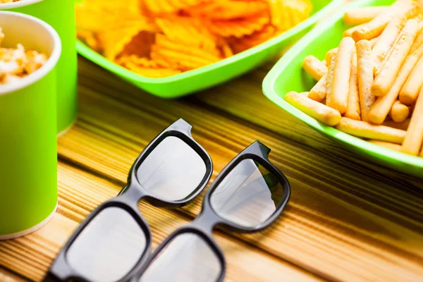 Hafta sonu evde, boş zaman tarzı, TV, fast food konsepti. 3D gözlükler, yeşil kaselerde uzaktan kumanda ve aperatifler: cips, patlamış mısır, ahşap arka planda hamburger. Üst Manzara. Kapat.