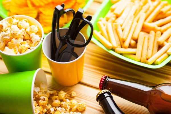 Hafta sonu evde, boş zaman tarzı, TV, fast food konsepti. 3D gözlükler, yeşil kaselerde uzaktan kumanda ve aperatifler: cips, patlamış mısır, ahşap arka planda hamburger. Üst Manzara. Kapat.