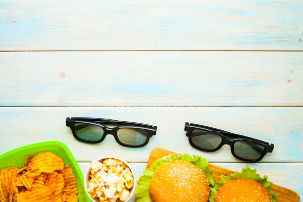 Hafta sonu evde, boş zaman tarzı, TV, fast food konsepti. 3D gözlükler, yeşil kaselerde uzaktan kumanda ve aperatifler: cips, patlamış mısır, ahşap arka planda hamburger. Üst Manzara. Kapat.