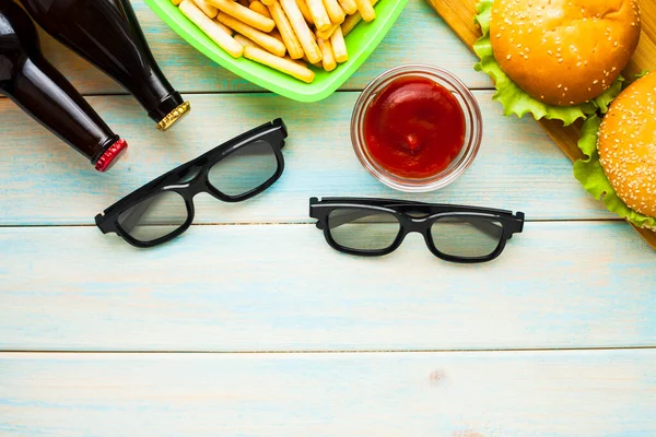 Hafta sonu evde, boş zaman tarzı, TV, fast food konsepti. 3D gözlükler, yeşil kaselerde uzaktan kumanda ve aperatifler: cips, patlamış mısır, ahşap arka planda hamburger. Üst Manzara. Kapat.
