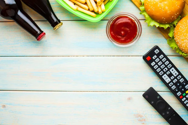 Hafta sonu evde, boş zaman tarzı, TV, fast food konsepti. 3D gözlükler, yeşil kaselerde uzaktan kumanda ve aperatifler: cips, patlamış mısır, ahşap arka planda hamburger. Üst Manzara. Kapat.