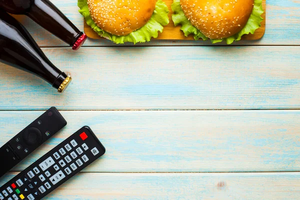 Hafta sonu evde, boş zaman tarzı, TV, fast food konsepti. 3D gözlükler, yeşil kaselerde uzaktan kumanda ve aperatifler: cips, patlamış mısır, ahşap arka planda hamburger. Üst Manzara. Kapat.