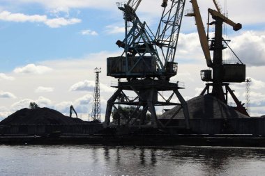 Mavi gökyüzüne karşı iskele vinçlerinin alt kısmının siluetleri (kabin; döner destek aygıtı; portal; yük arabaları; karşı ağırlık) ve yükleme için dağ kadar malzeme