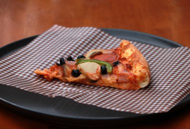 Kahverengi viski kağıdının üzerinde, tahta zemindeki siyah yuvarlak tabakta bir dilim pizzacı..