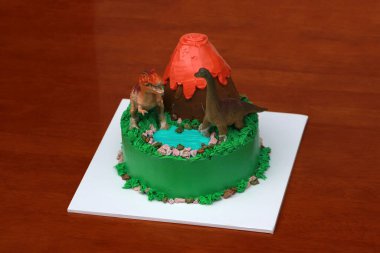 İki dinozor bebeği volkanik bir pasta ve ahşap zeminde beyaz kare bir tabakta yeşil bir orman yeri ile süslenir..