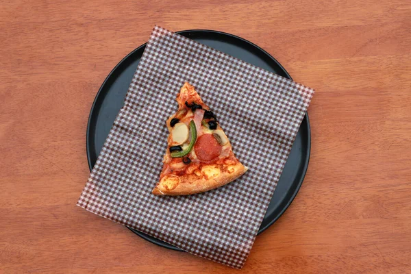 A piece of triangle Pizza  place on brown scotch paper  in the black round plate on the wooden floor.