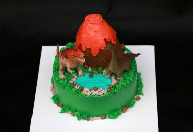 İki dinozor bebeği volkanik bir pasta ve yeşil ormanlık alanda siyah arka planda beyaz kare bir plaka üzerine dekore eder..