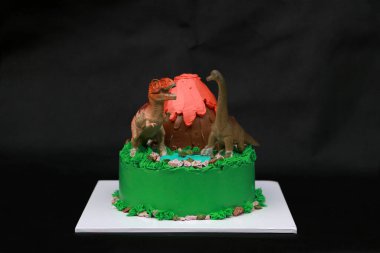 İki dinozor bebeği volkanik bir pasta ve yeşil ormanlık alanda siyah arka planda beyaz kare bir plaka üzerine dekore eder..