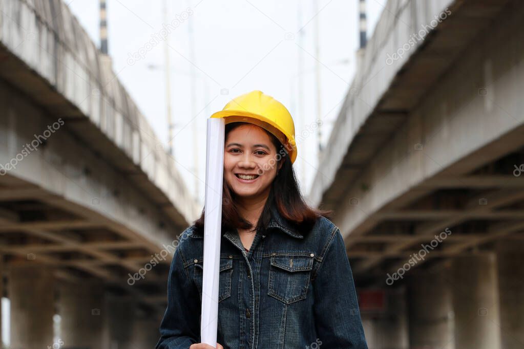 Female civil engineer or architect with yellow helmet, standing with project drafts while in hand on parallel expressway background.