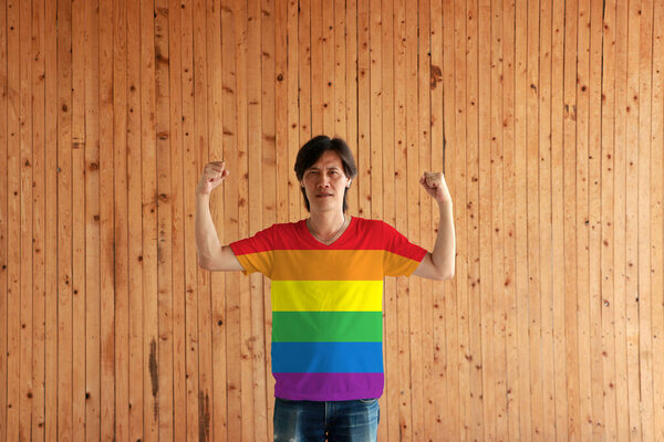 Man wearing symbol color of LGBTQ+ shirt and standing with raised both fist on the wooden wall background. The concept of lgbtq community.
