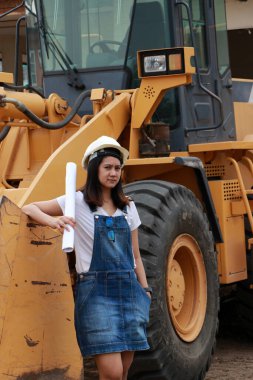 Beyaz miğferli ve proje taslaklı kadın inşaat mühendisi ya da mimar iş alanında Öndeki Yükleme Mühendisliği Araç Mühendisliği Aracı Alloy Buldozer Kamyonu ile birlikte dururken.