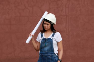 Beyaz miğferli kadın inşaat mühendisi ya da mimar elinde kırmızı kahverengi arka planda dururken proje taslakları.  