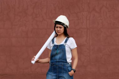 Beyaz miğferli kadın inşaat mühendisi ya da mimar elinde kırmızı kahverengi arka planda dururken proje taslakları.  