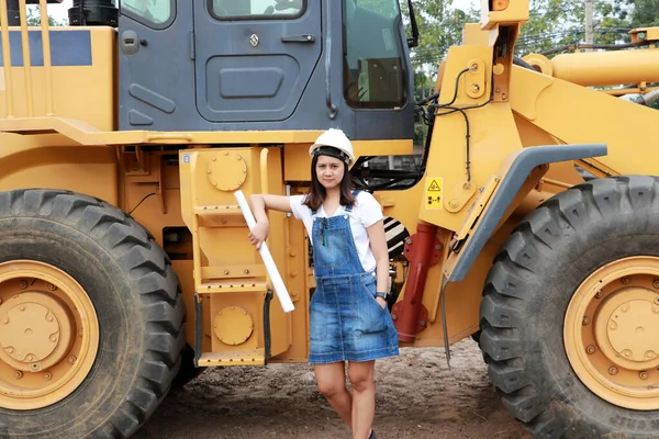 Female civil engineer or architect with white helmet and project drafts while in hand standing with Alloy Bulldozer Truck with Front Loader Truck Engineering Construction Car Vehicle at the work area.