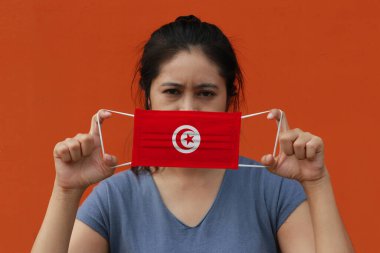 Elinde Tunus bayrağı, elinde hijyenik maskeyle turuncu arka planda ön yüzünü kaldırmış bir kadın. Minik parçacık ya da virüs koronası ya da Covid 19 koruması. Hastalıkla mücadele kavramı.