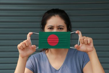 Elinde Bangladeş bayrağı, elinde hijyen maskesi olan ve ön yüzünü koyu yeşil arka planda kaldıran bir kadın. Minik parçacık ya da virüs koronası ya da Covid 19 koruması. Hastalıkla mücadele kavramı.