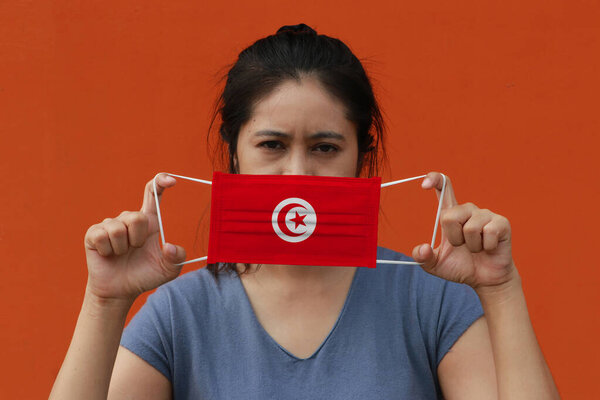 A woman with Tunisia flag on hygienic mask in her hand and lifted up the front face on orange color background. Tiny Particle or virus corona or Covid 19 protection. Concept of Combating illness.