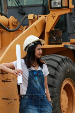 Beyaz miğferli ve proje taslaklı kadın inşaat mühendisi ya da mimar iş alanında Öndeki Yükleme Mühendisliği Araç Mühendisliği Aracı Alloy Buldozer Kamyonu ile birlikte dururken.