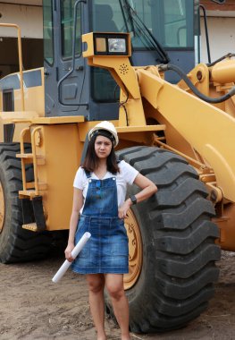 Beyaz miğferli ve proje taslaklı kadın inşaat mühendisi ya da mimar iş alanında Öndeki Yükleme Mühendisliği Araç Mühendisliği Aracı Alloy Buldozer Kamyonu ile birlikte dururken.