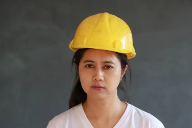 Kadın inşaat mühendisi ya da mimar sarı kasklı ve koyu gri arka planda beyaz tişört giyiyor.. 