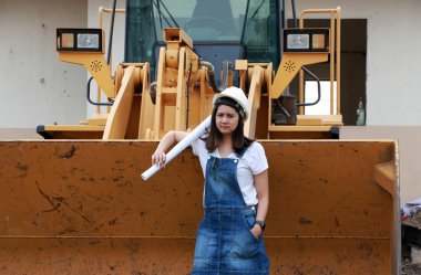 Beyaz miğferli ve proje taslaklı kadın inşaat mühendisi ya da mimar iş alanında Öndeki Yükleme Mühendisliği Araç Mühendisliği Aracı Alloy Buldozer Kamyonu ile birlikte dururken.